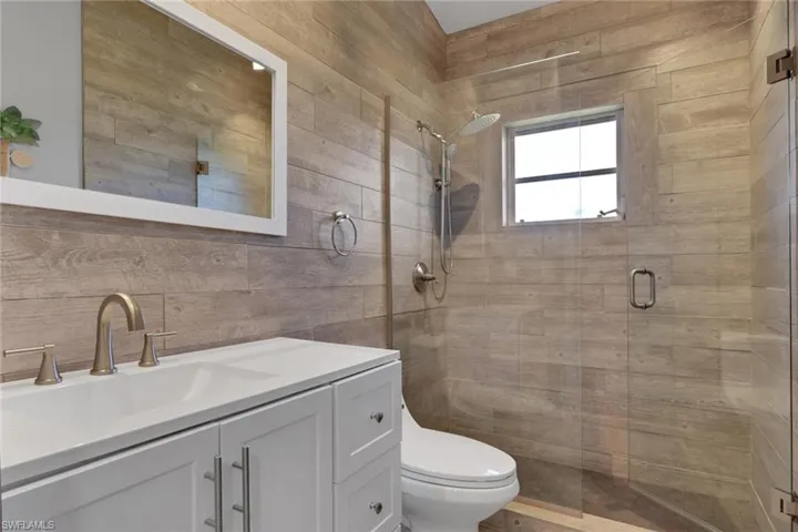 Full bathroom featuring a single white vanity with a rectangular undermount sink and brushed nickel faucet, wood-look tile wall accents, a glass-enclosed shower with a rain showerhead, and a casement window