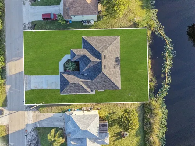Aerial perspective of a property featuring a multi-pitched shingled roof, a concrete driveway, and a grass-covered lot situated along a waterfront edge
