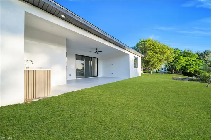 Covered patio featuring a built-in outdoor kitchen, vertical wood-slat cabinetry, ceiling fan, and recessed lighting, opening onto an expansive lawn