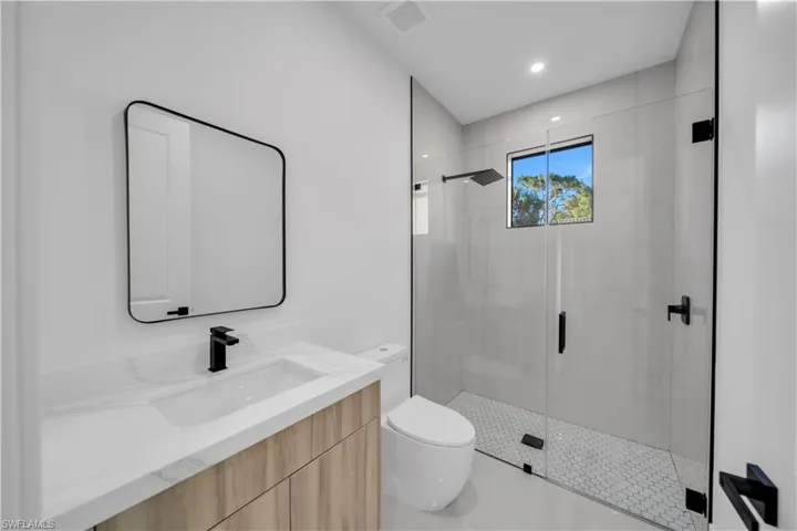 Bathroom featuring a wood-finish vanity with stone-surface countertop, matte black fixtures, a wall-mounted rectangular mirror, and a walk-in shower with a glass enclosure and hexagonal floor tile