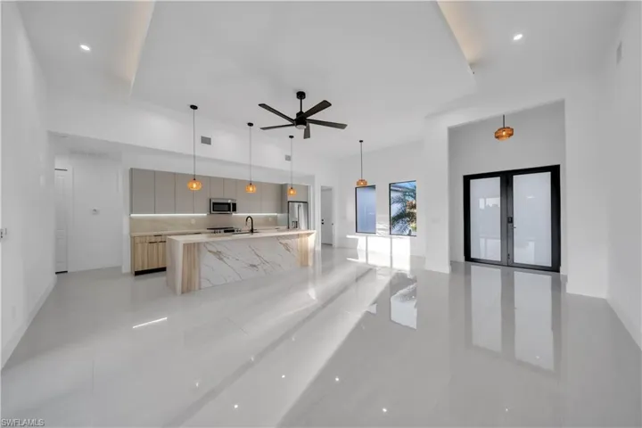 Open-concept kitchen featuring an oversized island with a waterfall-edge stone countertop, custom flat-panel cabinetry, recessed tray ceiling, large-format tile flooring, and matte black dual-pane exterior glass doors