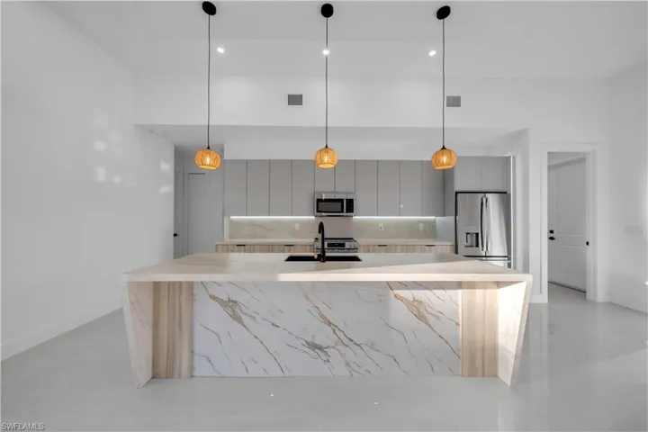 Kitchen featuring a waterfall-edge island with stone-patterned surfacing, flat-panel cabinetry, stainless steel appliances, under-cabinet lighting, and three suspended globe pendant lights