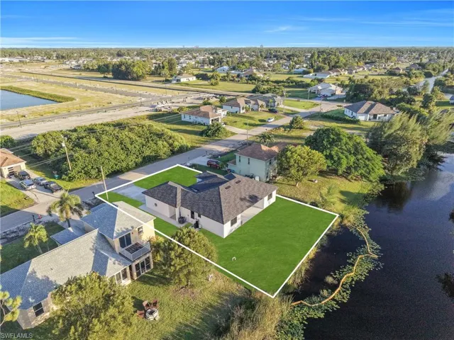 Aerial perspective of a single-story residence featuring a multi-pitched shingle roof, concrete driveway, and expansive lawn bordering a natural water feature