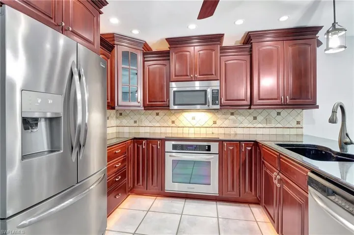 Kitchen with stainless steel appliances, bold wood finish cabinets, dark stone countertops, glass fronted cabinets, and decorative light fixtures.