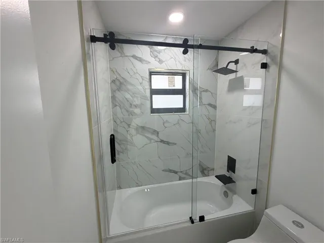 Bathtub with a glass sliding shower door featuring matte black hardware, white marble-patterned wall tile, a built-in shower niche, and a contrasting matte black shower head