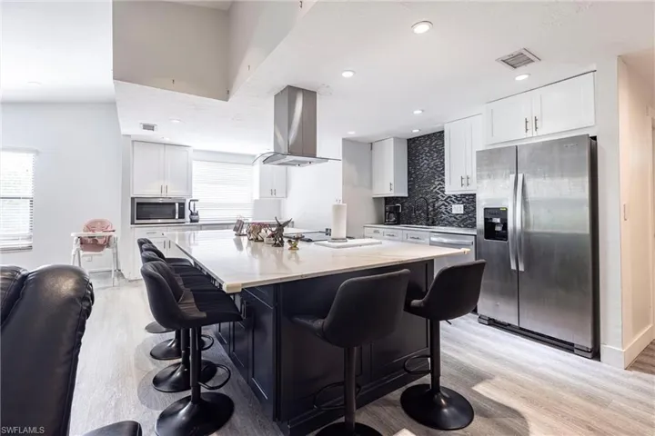 Kitchen with appliances with stainless steel finishes, backsplash, light stone countertops, light wood-style flooring, and island range hood