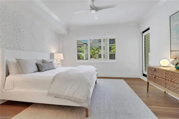Large primary bedroom featuring wood floors, Serena & Lily wallpeper, plantation shutters, access to the balcony, and ceiling fan