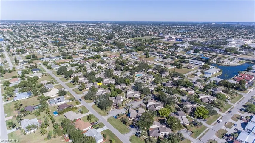 Aerial view of a residential area featuring numerous single-story homes, mature landscaping, paved streets, and a prominent body of water in the distance