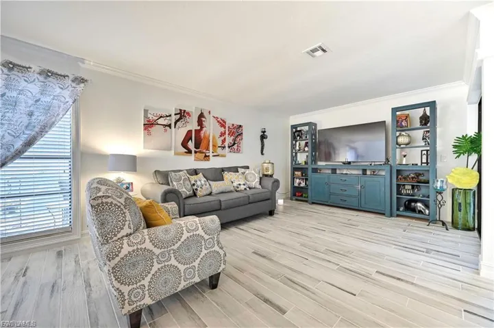 Spacious living area featuring light-toned wood-finish flooring, a large window with blinds, substantial built-in entertainment cabinetry, and crown molding