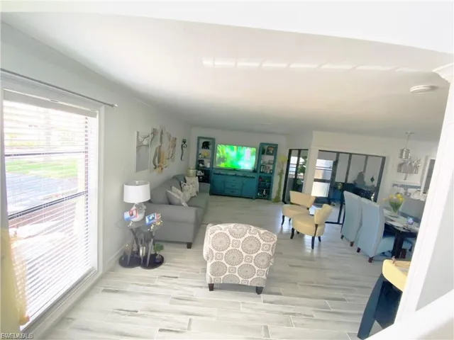 Open-concept living space with light-toned wood-finish flooring, a large window with blinds, and white walls