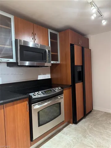 Kitchen featuring wood-finish cabinetry with some frosted glass door fronts, black countertops, and stainless steel appliances including an oven range and microwave