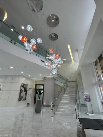 Grand foyer featuring a multi-level chandelier with clear and amber glass globes, a marble staircase with glass and stainless steel railings, polished tile flooring, and high ceilings
