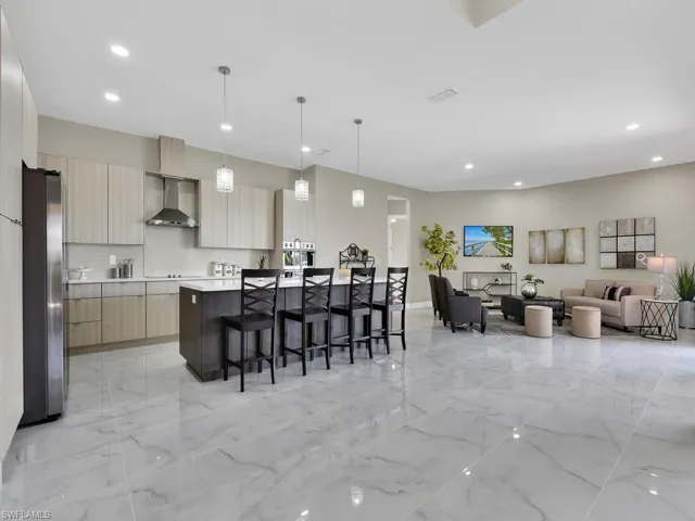 Kitchen featuring light brown cabinets, hanging light fixtures, wall chimney exhaust hood, a kitchen island with sink, and stainless steel refrigerator