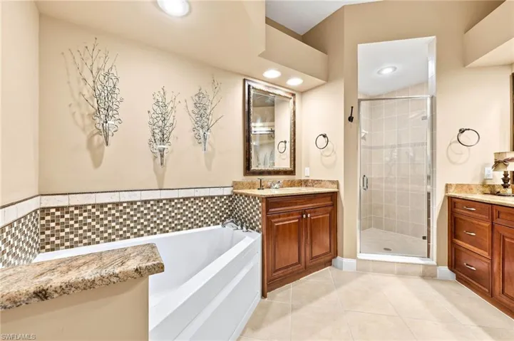 Spa-inspired primary bathroom featuring a soaking tub with decorative tile surround, dual wood-finish vanities with granite countertops, a frameless glass-enclosed shower with tile detailing, recessed lighting, and decorative wall art