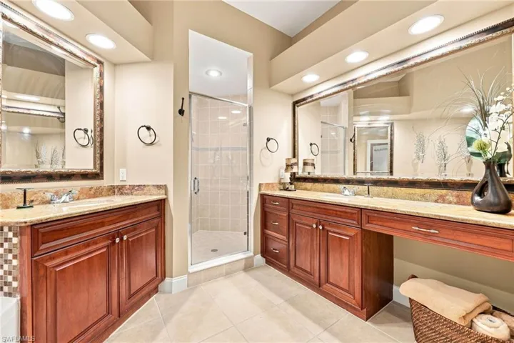 Luxurious primary bathroom featuring dual vanities with wood-finish cabinetry, granite countertops, and framed mirrors with integrated lighting