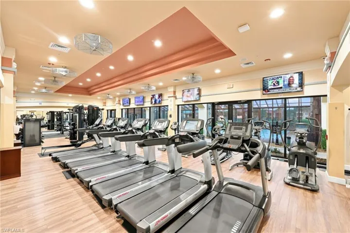 State-of-the-art fitness facility featuring multiple treadmills with individual entertainment displays, comprehensive strength training equipment, wood-finish flooring throughout, recessed and pendant lighting, and expansive windows providing natural light and outdoor visibility