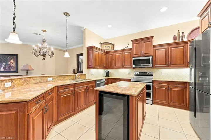 Spacious kitchen featuring rich wood cabinetry, granite countertops, a large center island with black base, stainless steel appliances including a gas range and refrigerator, pendant lighting, and ceramic tile flooring