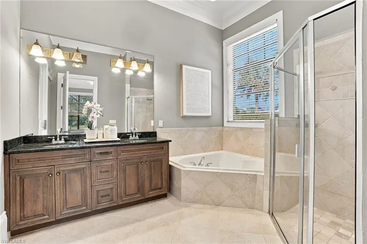 Dual-sink vanity with dark stone countertop, large wall-mounted mirror, corner soaking tub, and walk-in shower with tiled surround