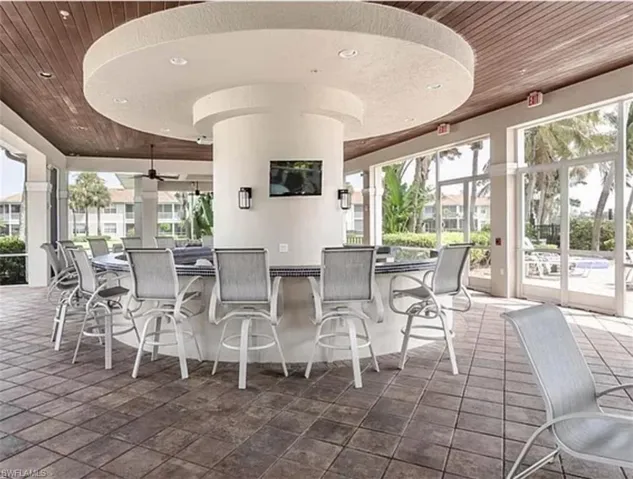 Expansive covered outdoor living area featuring a central bar with seating, wood-finish ceiling, tiled flooring, and large glass panel doors