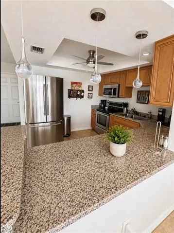 Kitchen featuring granite countertops, stainless steel appliances, wood cabinetry, contemporary pendant lighting, and a recessed ceiling with a ceiling fan