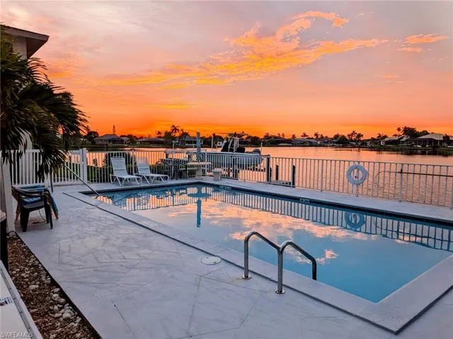 Waterside property featuring a rectangular pool with integrated steps, a large paved pool deck, and a white picket fence bordering the canal