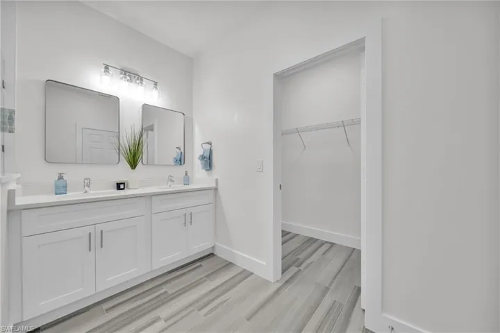 Dual-sink vanity with white cabinetry, light-toned countertop, rectangular undermount basins, two wall-mounted mirrors, and multi-bulb vanity lighting, complemented by tile flooring and an integrated walk-in closet with wire shelving