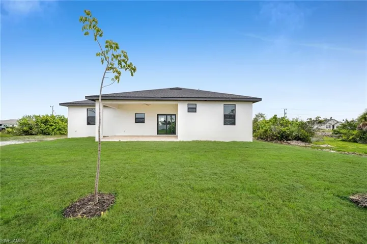Rear exterior featuring a white stucco facade, a covered patio with a ceiling fan, a hip roof with asphalt shingles, and expansive open green space