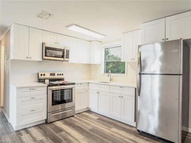 Kitchen featuring white shaker-style cabinetry, stainless steel appliances, light-toned countertops, wood-finish flooring, and a window above the sink