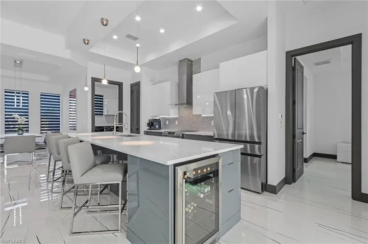 Kitchen featuring stainless steel appliances, wall chimney range hood, decorative light fixtures, beverage cooler, and a raised ceiling