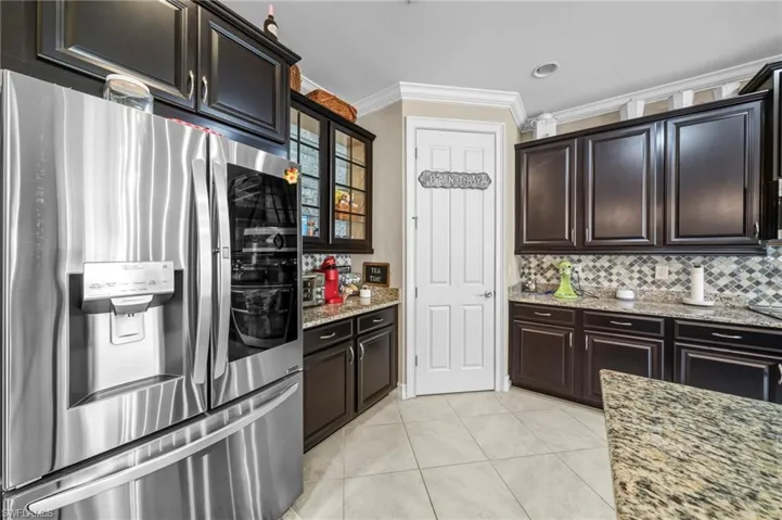 Kitchen with stainless steel fridge, ornamental molding, light tile patterned floors, light stone counters, and glass insert cabinets