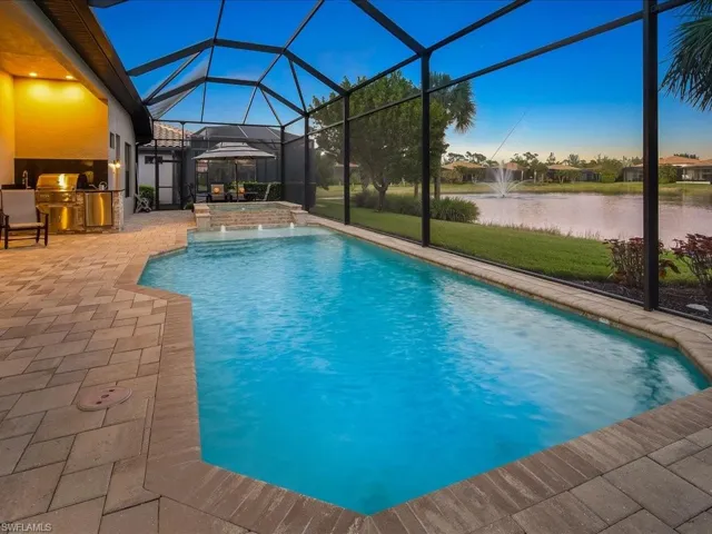 Pool at dusk with a lanai, pool water feature, exterior kitchen, a water view, and a patio area