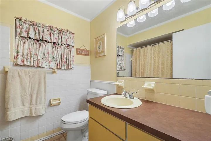 Bathroom featuring a single vanity with an integrated sink, a rectangular mirror with an overhead light fixture, a toilet, and a tiled shower with a shower curtain