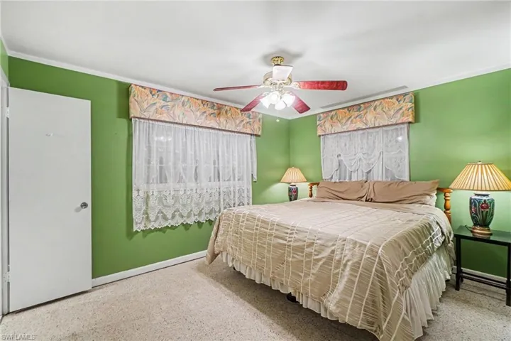 Carpeted bedroom featuring a ceiling fan with integrated lighting, two windows with valances and lace curtains, and a white door with a round knob