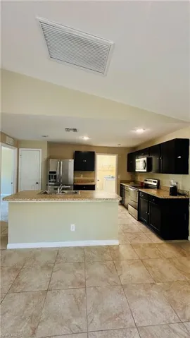 Kitchen featuring dark cabinets, stainless steel appliances, an island with sink, light stone countertops, and washer / dryer