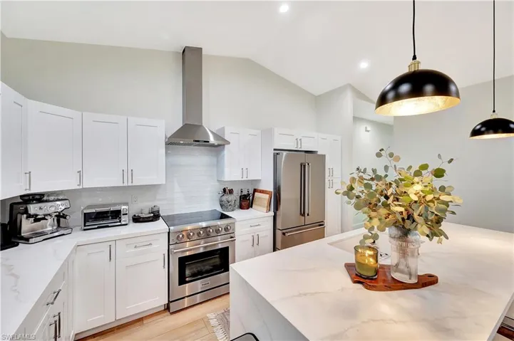 Kitchen with wall chimney range hood, high quality appliances, vaulted ceiling, decorative light fixtures, and white cabinetry
