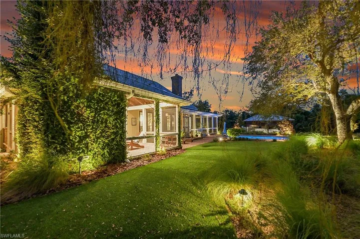 Twilight view of the beautifully landscaped grounds, where soft landscape lighting frames the lawn and highlights the covered outdoor living area with fireplace, offering an elegant space for evening entertaining