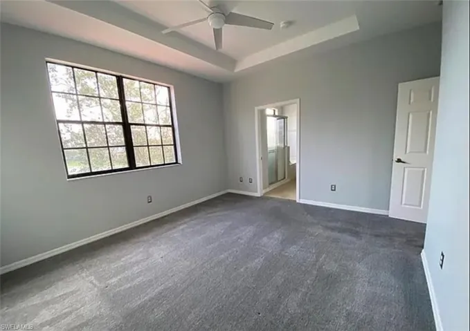 Unfurnished bedroom featuring dark colored carpet, a tray ceiling, ensuite bath, and ceiling fan