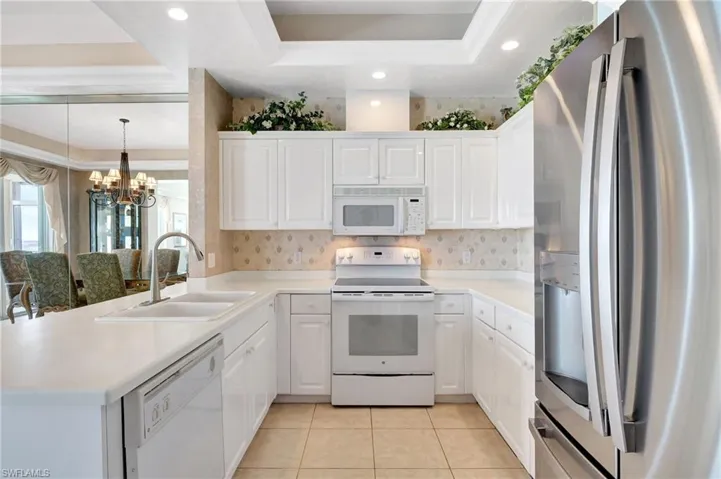 Kitchen featuring white appliances, light tile patterned floors, a raised ceiling, a sink, and a notable chandelier