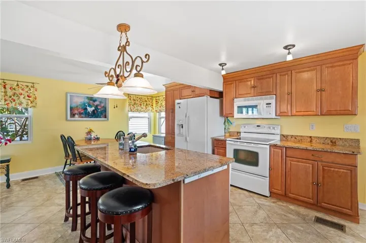 Kitchen with white appliances, hanging light fixtures, an island with sink, and granite counters