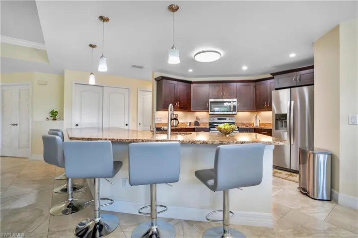 Kitchen with stainless steel appliances, light stone countertops, dark wood finish cabinets, a center island with sink, and pendant lighting