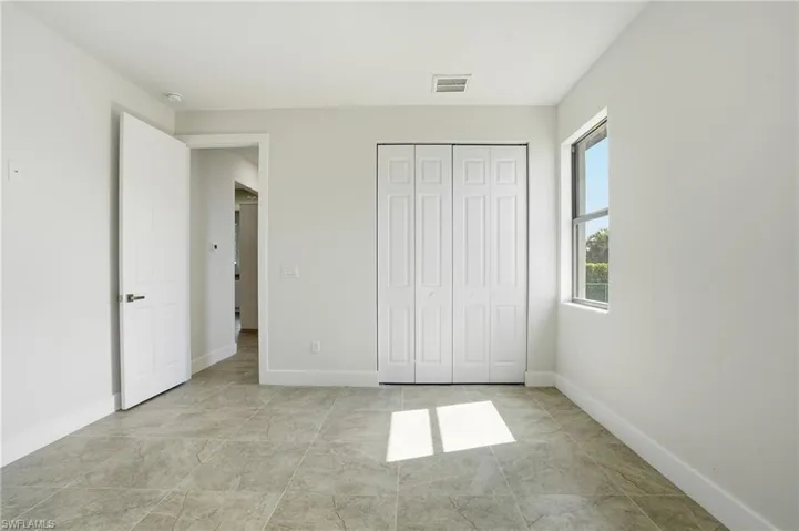 Neutral-toned bedroom featuring large format tile flooring, a white bi-fold closet, a white panel door with dark hardware, a single window, and white baseboards
