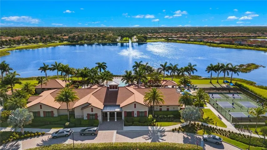 Expansive community recreation building with a large lake backdrop, featuring multiple pickleball courts, a central fountain, and lush landscaping with mature palm trees