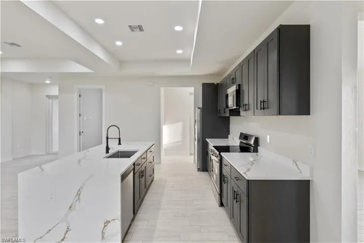 Kitchen featuring stainless steel appliances, light stone countertops, an island with sink, recessed lighting, and a raised ceiling