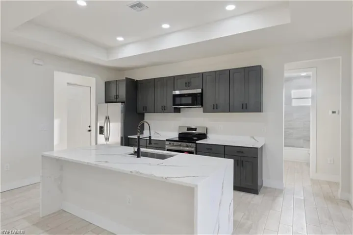 Kitchen featuring stainless steel appliances, a raised ceiling, light stone countertops, light wood finished floors, and an island with sink