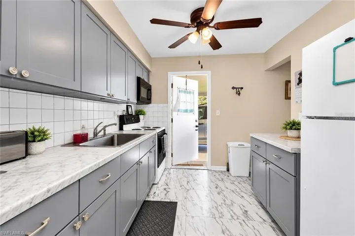Galley kitchen featuring gray cabinetry with hardware accents, marble-finish countertops, stainless steel sink and range, white subway tile backsplash, ceiling fan with integrated lighting, and stone-look tile flooring throughout