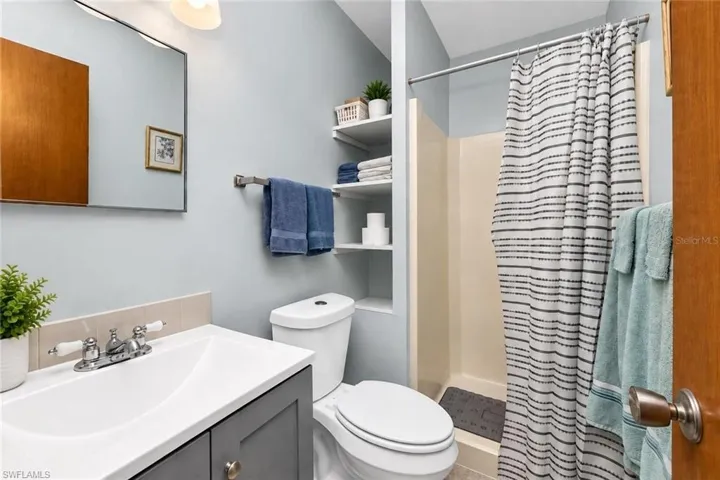 Vanity with white sink and gray cabinetry, white toilet, built-in shelving, and a shower with a curtain rod