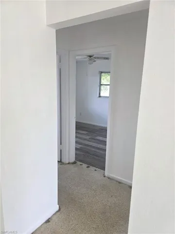 Interior hallway with light-colored carpeting, leading to a room featuring wood-finish flooring, a window, and a ceiling fan