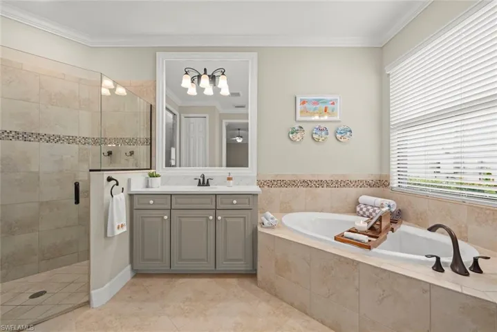Primary bathroom, two vanities, tiled flooring and walls, a glass-enclosed shower, a built-in soaking tub with dark bronze fixtures, and a vanity with a white countertop and gray cabinetry