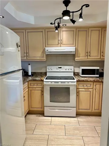 Kitchen with white appliances, under cabinet range hood, backsplash, and dark stone counters