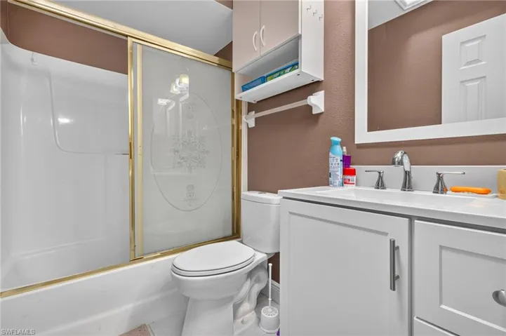 Guest bathroom featuring a white vanity with a white countertop, chrome faucet, wall-mounted mirror, and a combined tub and shower unit with a gold-framed frosted glass enclosure